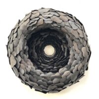 The sculpture "New Moon" features layered, overlapping dark metal discs arranged in a circular, tunnel-like formation against a white background.